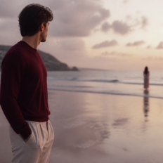 An adult stands alone by reflective water, looking toward a distant blurred figure.